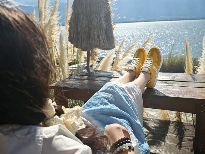 A woman relaxes on a wooden deck overlooking a sparkling body of water, her feet propped up in yellow sneakers and her head resting against a post.