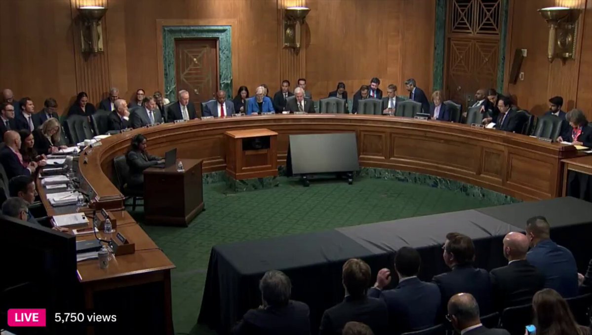 The image shows a group of people sitting around a large wooden table, with some individuals seated at the head of the table while others are scattered across its surface. The scene appears to be taking place inside a courtroom or a similar setting where a meeting is being held. A TV screen can be seen in front of the attendees, possibly displaying information relevant to their discussion.

The people present seem to be engaged in a serious conversation, with some individuals wearing ties as part of their attire. The room has wooden walls and chairs arranged around the table, creating an atmosphere of formality and professionalism.