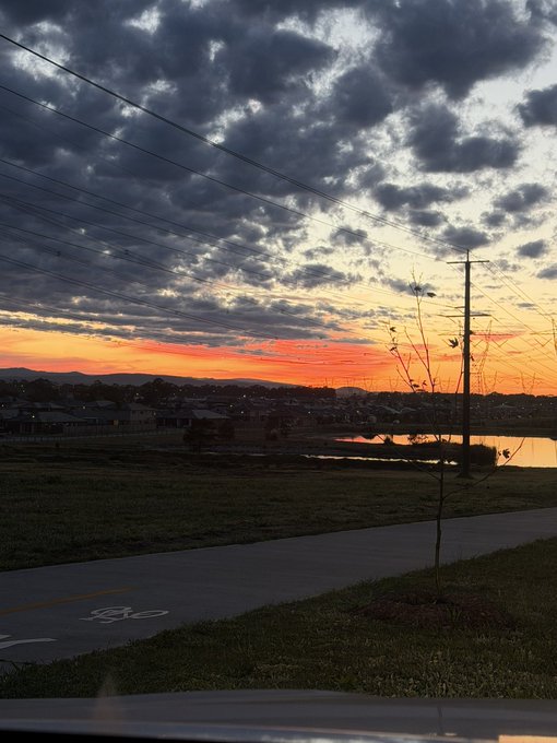 A scenic sunset photograph taken from a car, showing dramatic cloud-filled skies over a pond and suburban landscape with power lines and a small tree silhouetted in the foreground.