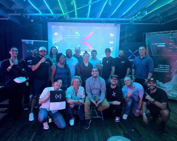 A large group of people pose together at what appears to be a tech event, with "XRP LEDGER" branding visible on the backdrop and attendees wearing branded clothing.