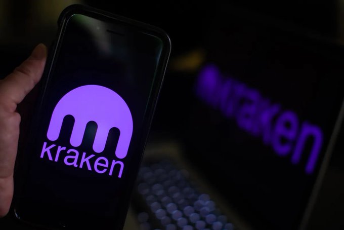 A hand holding a smartphone displaying the purple Kraken cryptocurrency exchange logo, with a blurred monitor showing the same logo in the background.