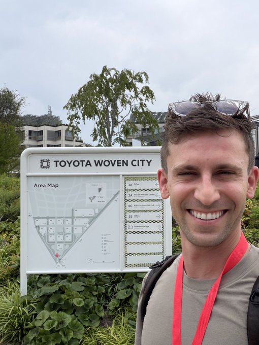A selfie of a smiling man with sunglasses on his head standing in front of a Toyota Woven City area map sign with greenery and buildings in the background.