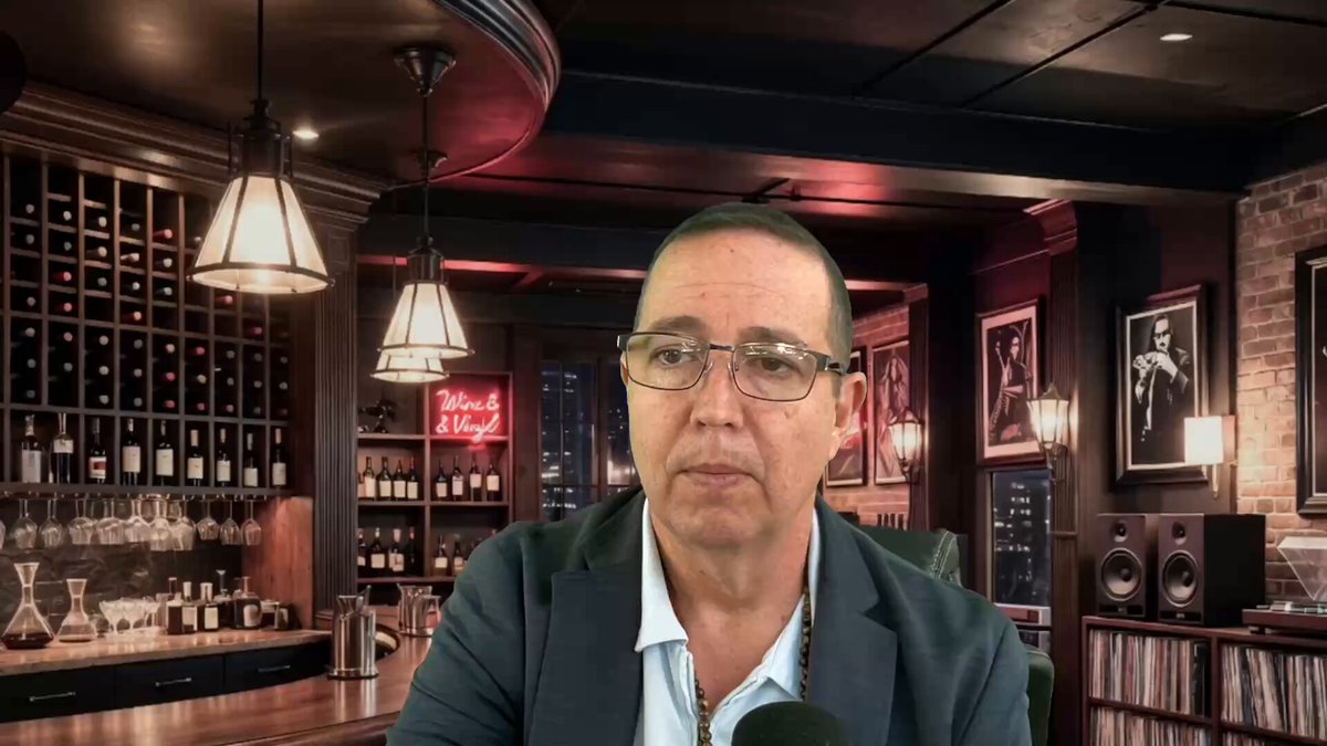 The image features a man sitting at a bar, wearing glasses and a suit jacket. He appears to be talking into a microphone or headset, possibly recording a podcast or live stream. The background of the image shows a wine cellar with numerous bottles displayed on shelves. There is also a neon sign in the room that reads "Crypto Bloodbath Of XRP?".