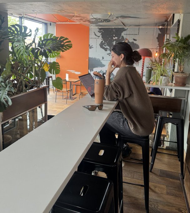 A woman sits at a white counter in a stylish café, working on a laptop with a water bottle and sunglasses beside her, surrounded by lush green plants and warm orange walls.