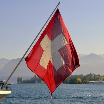 A Swiss flag waves against a backdrop of mountains and a lake, symbolizing the nine Swiss firms piloting the CHFD stablecoin to test programmable payment solutions domestically.