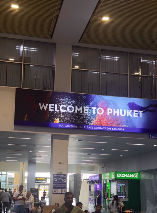 A traveler stands beneath a large illuminated "WELCOME TO PHUKET" banner at the airport arrivals hall, with a green EXCHANGE booth and other travelers visible nearby. The image captures the start of the poster's Thailand trip in Phuket, matching the "#Phuket" tag in the caption.