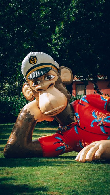 An inflatable monkey wearing a white captain's hat and a red patterned shirt lies on the grass, resting its chin on its hand in a thoughtful pose.