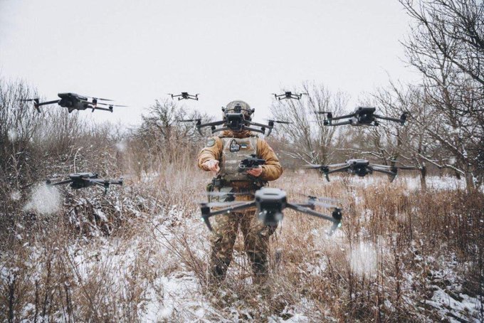 A soldier in camouflage walking through a snowy field surrounded by multiple hovering drones.