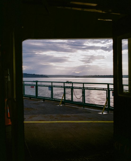 A scenic view through an open ferry doorway, framing calm water, distant mountains, and a cloudy sky at dusk with a green safety railing in the foreground.