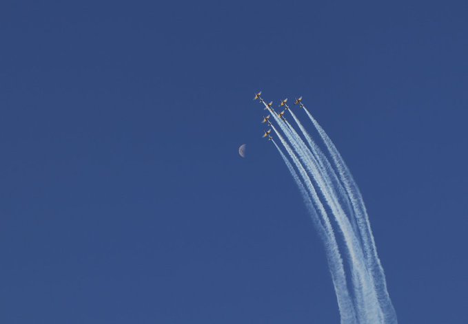 A formation of jet aircraft flying upward against a clear blue sky, leaving contrails alongside a visible crescent moon.