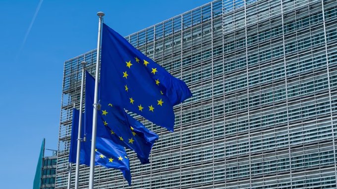 Three European Union flags waving in the wind in front of a modern glass office building under a clear blue sky.