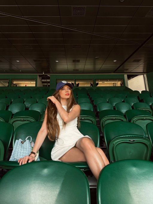 A woman in a white dress and blue baseball cap sits in green stadium seats with a light blue handbag beside her.