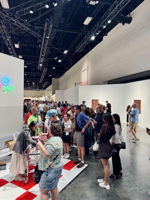 A crowded art exhibition or fair with many attendees mingling in a large hall with white walls and industrial ceiling lighting.