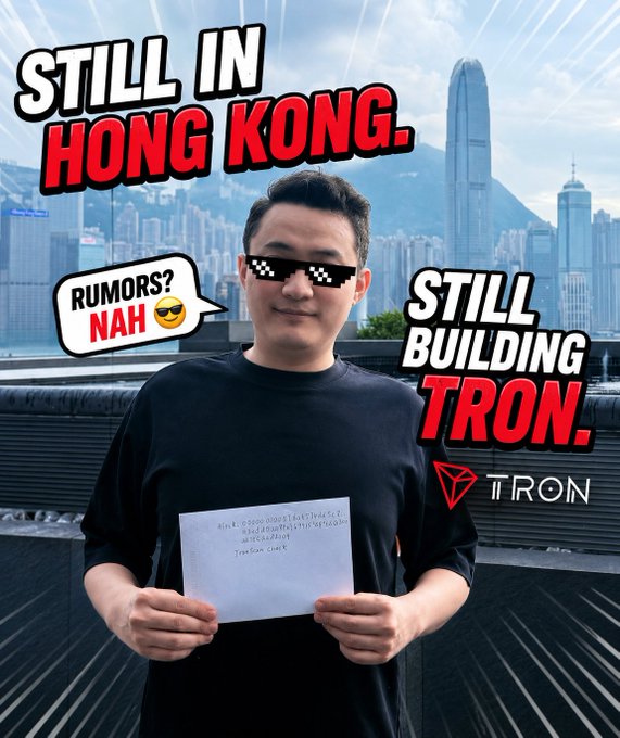 Justin Sun poses in Hong Kong with the Hong Kong skyline behind him, holding up a handwritten note (dated 2026) referencing a TRON mainnet checkpoint, while bold text reads "STILL IN HONG KONG" and "STILL BUILDING TRON," alongside the TRON logo and a "RUMORS? NAH 😎" speech bubble—visually debunking rumors that he left the city.