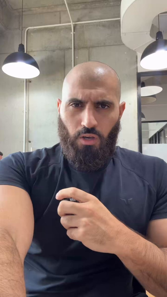 The image features a man with a beard wearing a black shirt, sitting at a desk or table. The man has his arms crossed and appears to be deep in thought. There are two people visible in the background of the photo, but they do not seem to be interacting with the main subject.