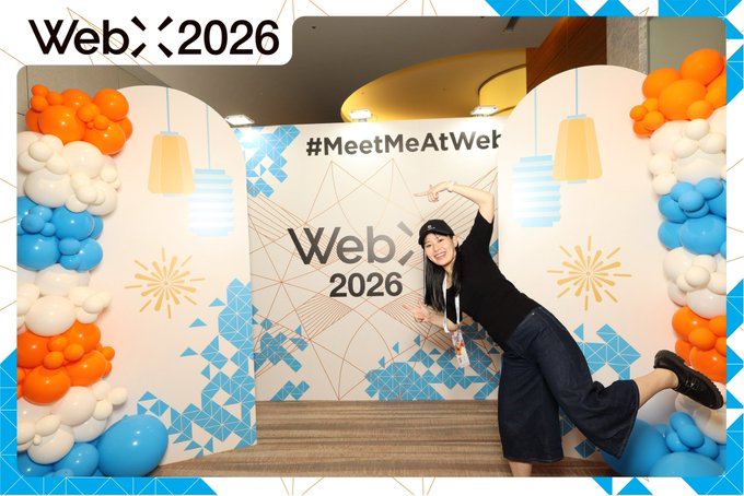 A woman poses playfully at a 'Web> 2026' conference booth decorated with blue and orange balloons and the hashtag '#MeetMeAtWeb'.