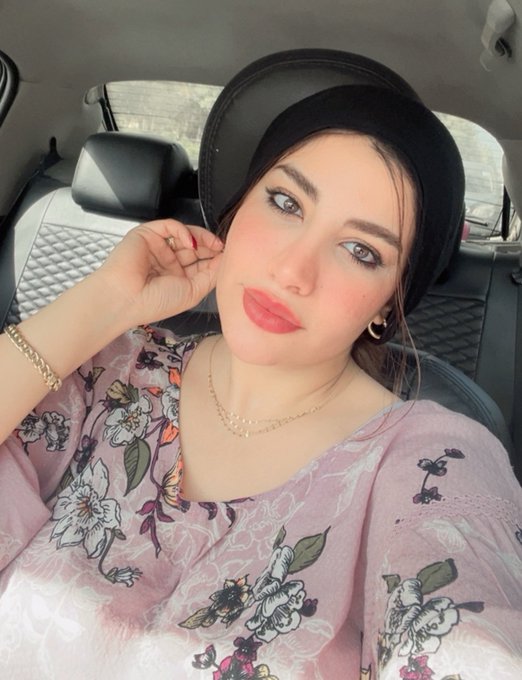 A selfie of a woman wearing a black hijab and a pink floral top, sitting in a car with quilted black leather seats.