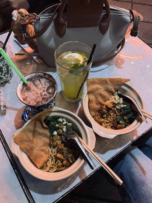A tabletop scene showing two bowls of noodles with crispy crepes and greens, accompanied by a lemon-mint drink in a tall glass, an iced coffee, and a grey handbag in the background.