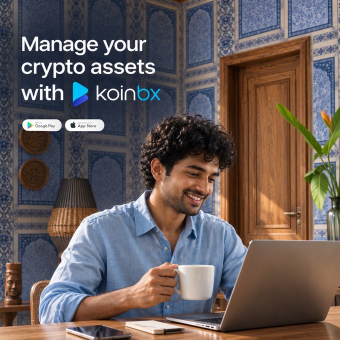 A young man with curly hair smiles while holding a coffee mug at a laptop in a room with ornate blue patterned walls and a wooden door. The advertisement reads "Manage your crypto assets with koinbx," featuring Google Play and App Store badges, promoting a reliable crypto management platform that aligns with the post's message about prioritizing trustworthy, user-friendly crypto services for India's evolving market.