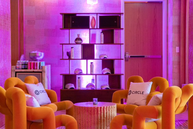 Two orange tubular chairs with "CIRCLE" branded pillows flank a wooden stump coffee table in a lounge illuminated by pink and purple lighting, with a shelving unit of decorative items in the background. The branded pillows visually tie the cozy, branded space to Circle's presence at Sibos 2026 in Miami, where the company engaged in conversations about the future of digital financial infrastructure.