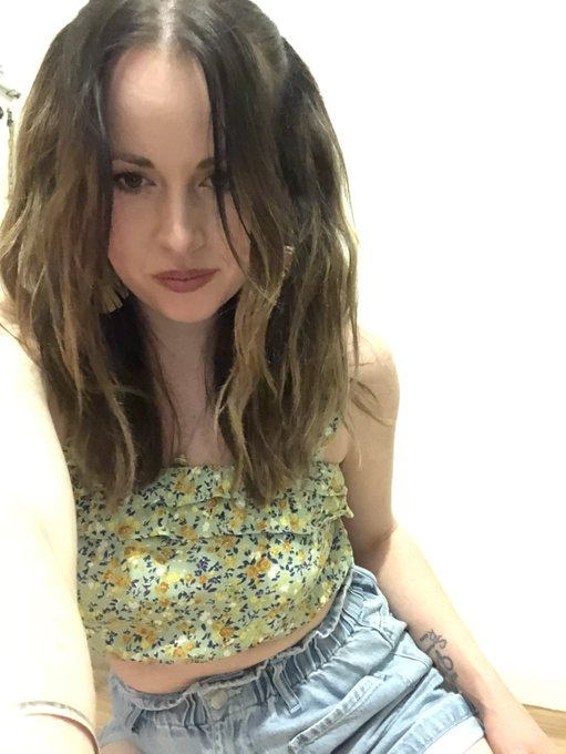 A selfie-style photo of a woman with long wavy dark hair, a floral yellow top, and light denim shorts, with a small tattoo visible on her wrist.