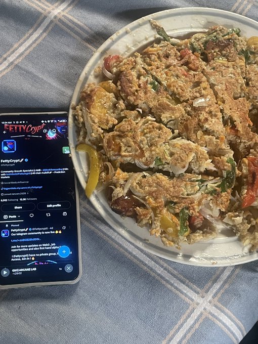 A hearty plate of scrambled eggs with peppers and onions sits beside a smartphone displaying the FettyCrypt Twitter/X profile with its "FettyCryptCommunity" community and related crypto hashtags, showing the user enjoying their breakfast while engaging with their online community.