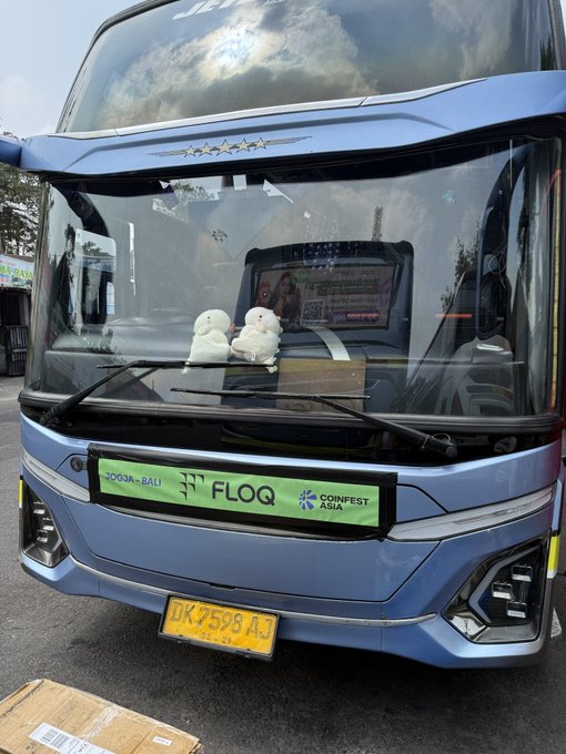 The front of a blue bus with 'JOGJA - BALI', 'FLOQ', and 'CONFEST ASIA' signage, featuring two small plush toys on the dashboard.