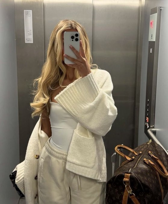 A woman with long blonde hair takes a mirror selfie in an elevator while wearing a white tube top, cream knit cardigan, and beige sweatpants, posing with a Louis Vuitton Speedy monogram bag beside her. The "gm ct" caption (good morning Connecticut) pairs with the casual, stylish morning look captured in the metallic elevator setting.