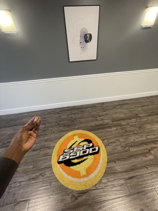 First-person view of a hand near an orange and yellow Bitcoin-themed rug on a wooden floor, with framed art on a gray wall behind it.