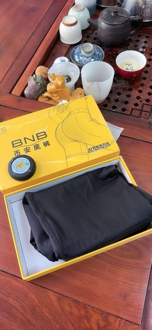 An open yellow box labeled 'BNB' and '币多足裤' (Binance underwear) sits on a wooden tea table beside a teapot and teacups, displaying a folded black garment inside.