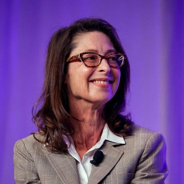 A smiling woman with shoulder-length brown hair and glasses, wearing a gray blazer over a white collared shirt, speaks at an event against a purple backdrop, likely representing Fidelity CEO Abigail Johnson expressing her positive stance on Bitcoin as a savings asset.