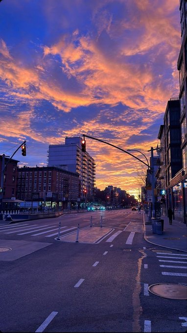 A vibrant urban sunset paints the sky in fiery oranges and pinks above a quiet city street intersection, with traffic lights glowing and buildings silhouetted against the dramatic clouds. The caption "GM if you GM" suggests this breathtaking morning view is a greeting to fellow crypto or social media users who respond with their own "good morning" ("GM").