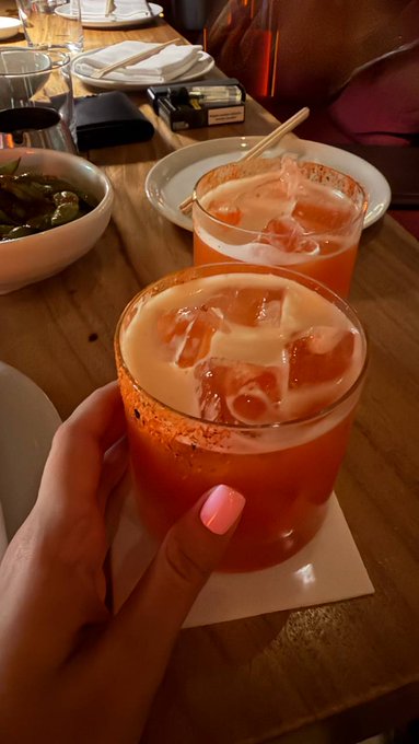 A close-up of a hand with pink nail polish holding an orange cocktail with ice and a chili-salt rim on a coaster, with another similar drink and a bowl of edamame on the table.