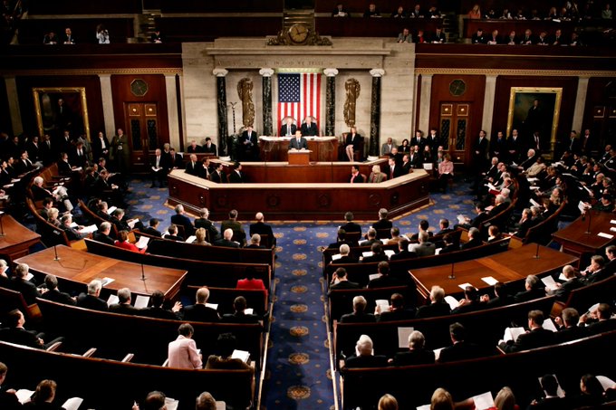 A wide view of the U.S. House of Representatives chamber during a session, with members seated in the curved desks and an American flag displayed at the front.