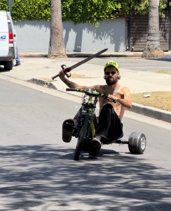 A shirtless man wearing a yellow hat with "SKOOT" visible rides a green drift trike while holding a sword, embodying a wild spirit for "SOLANA SUMMER."