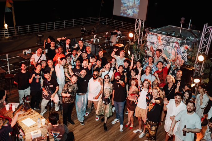 A large group of people poses together on the deck of a boat at night, with tropical decor, lanterns, and a projection screen visible in the background.