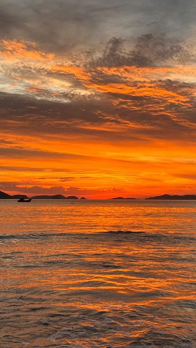 A vibrant orange and yellow sunset over the ocean with silhouettes of islands and a boat on the horizon.