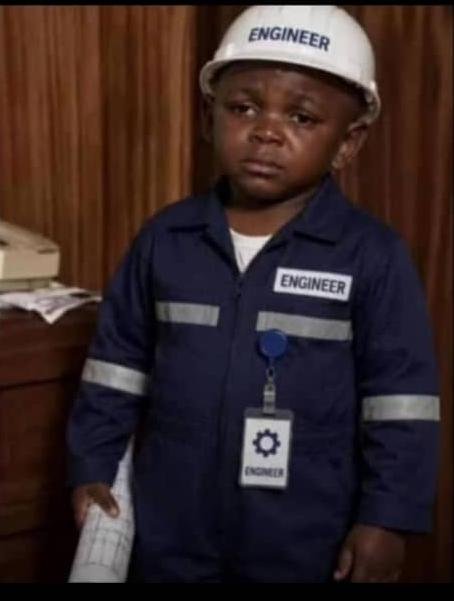 A photograph of a young child wearing a navy blue work uniform and white hard hat, both labeled 'ENGINEER,' with an ID badge, holding a rolled-up paper, appearing to be a costumed scene from a television show.