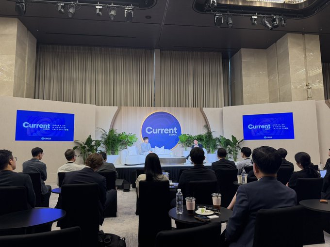 A conference or panel discussion at a 'Current' branded event, with two speakers on stage in front of screens displaying the Current logo, while an audience in business attire watches from seated tables.