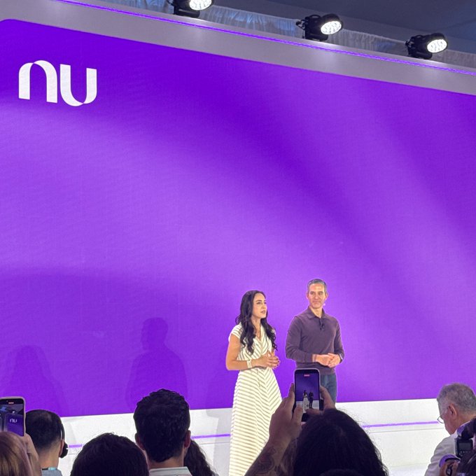Two presenters stand on a stage in front of a large purple backdrop featuring the 'nu' logo, likely at a Nubank event.