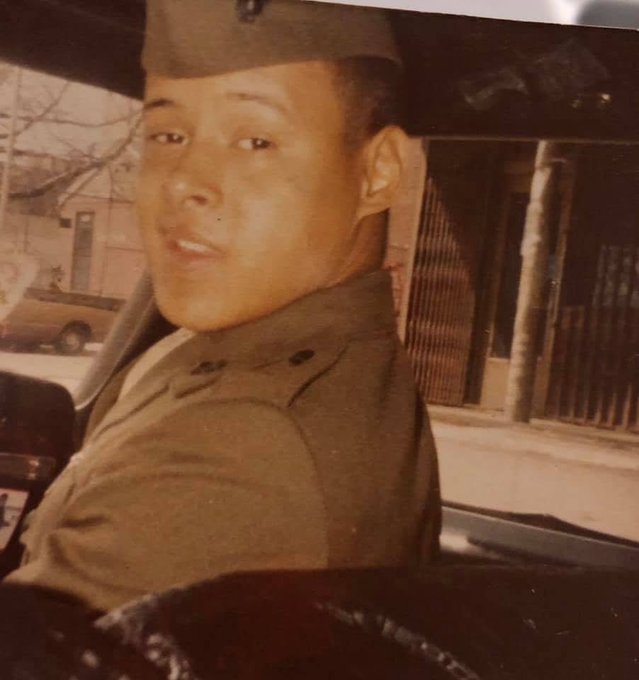 A vintage photograph of a young man in a military uniform and garrison cap, looking over his shoulder from inside a vehicle, with a street and car visible behind him.