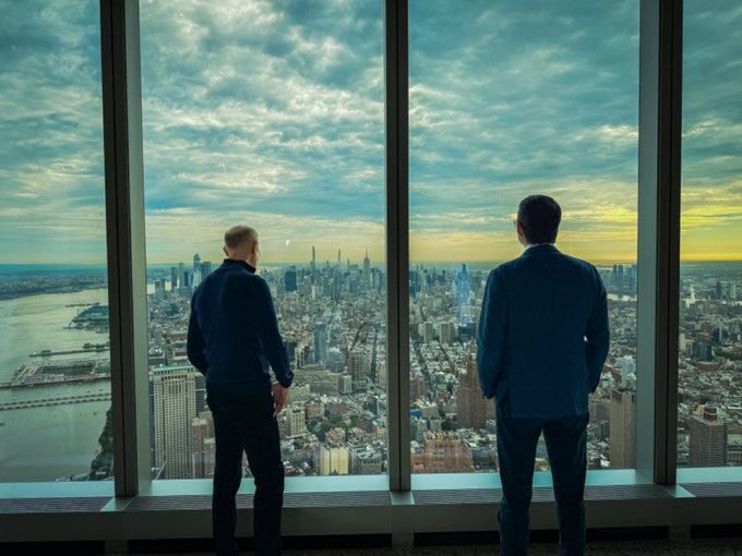 Two men in suits stand with their backs to the camera, looking out a large window at a sprawling city skyline, likely New York, at sunset.