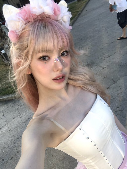 A young woman with pastel pink hair styled in twin tails poses outdoors, wearing a white corset top and a flower crown headband adorned with cat ears, matching the K-pop idol aesthetic tagged in the post. The hashtags "#KiiiKiii #키키 #SUI #수이" suggest this is a fan photo or official content featuring Sui, a member of the K-pop group KiiiKiii.