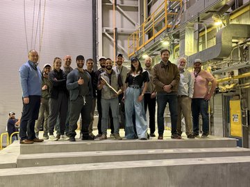 The image shows a group of people standing together, posing for a photo. They are all dressed similarly, wearing dark clothing that contrasts with their surroundings. The setting appears to be an industrial or warehouse environment, as evidenced by the presence of pipes and other equipment in the background. A yellow door can also be seen in the scene. The post caption reads: "this is so awesome! congratulations to the entire team!!". This message seems to be a congratulatory note for the group's achievement or success, possibly related to their work at the industrial facility.