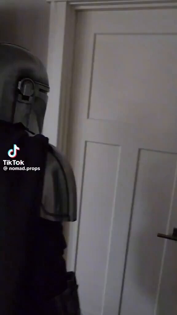 The image features a person wearing a black outfit with a helmet or armor-like suit, standing next to an open white door. The person appears to be shaking their body due to the excitement of being in front of the camera. There are also some text elements present in the image that read " TikTok 3D props".