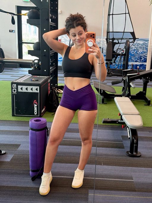 A woman takes a gym mirror selfie in a black sports bra and purple shorts, posing with a yoga mat nearby. The post's "looksmaxxing" theme is highlighted by the workout gear and fitness equipment in the background, aligning with the BTC-themed tagline about building both stacks and aesthetics.