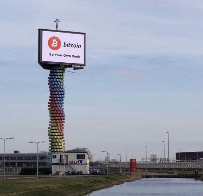 A tall tower of stacked books displays a sign with the Bitcoin logo and text 'bitcoin Be Your Own Bank' next to a canal.