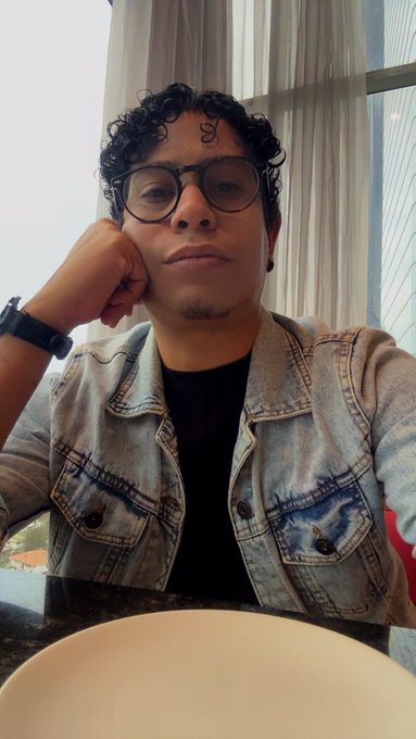A young man with curly hair, glasses, and a denim jacket rests his head on his hand while sitting indoors near a window.