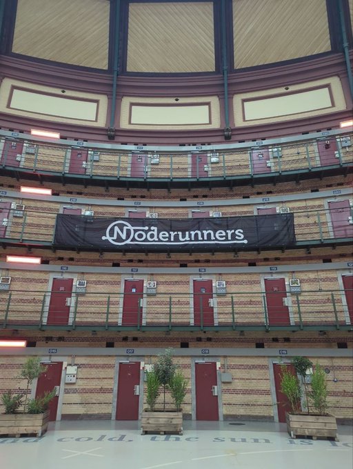An interior view of a circular prison-style structure with multiple tiers of doors, featuring a black banner reading "Noderunners" hanging across the center, with plants arranged at the bottom.