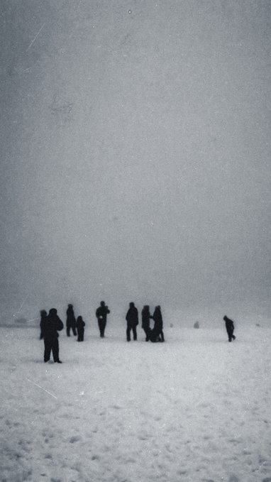 A grainy, atmospheric photograph showing silhouettes of several people standing in a vast, foggy snow-covered landscape under a gray sky.